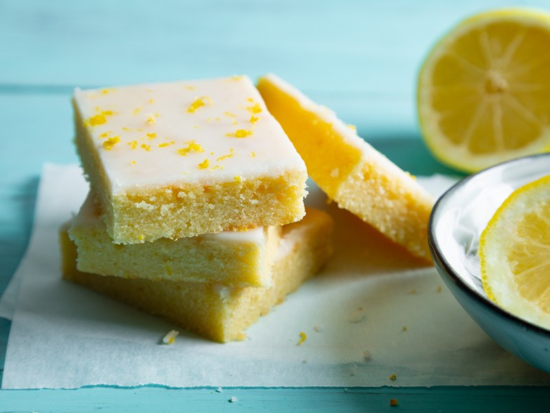 4 Stücke Zitronen-Brownies Lemonies auf Backpapier, daneben halbierte Zitronen.