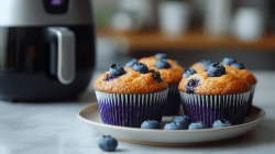 ein paar Blaubeermuffins aus der Heißluftfritteuse auf einem Teller, im Hintergrund unscharf eine solche Fritteuse.