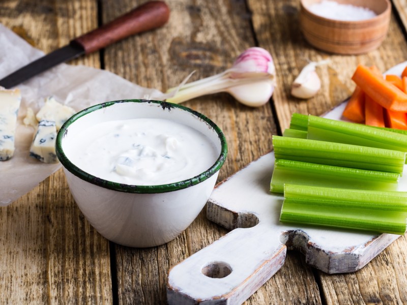 Eine Schale Blauschimmelkäse-Dressing, daneben Gemüse-Sticks und ein angeschnittener Köse sowie Knoblauch.