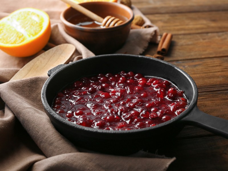 Cranberry-Kompott in einer Pfanne, daneben eine halbe Orange und eine Schale Honig.