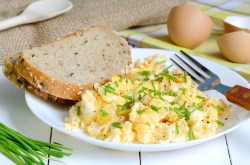 Ein Teller Rührei mit Crème fraîche und zwei Scheiben Brot, daneben Schnittlauch und Eierschalen sowie eine Gabel.