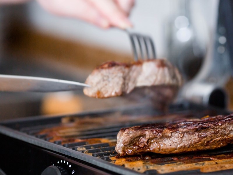 Amazon Black Friday Tefal OptiGrill Kontaktgrill: 2 Stück Fleisch auf einem Kontaktgrill, eins wird mit Messer und Gabel weggenommen. Nahaufnahme.