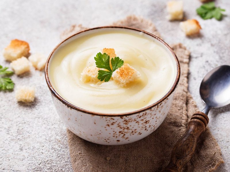 Eine Schüssel Apfel-Blumenkohlsuppe mit Croûtons, drumherum weitere Croûtons und ein Löffel.