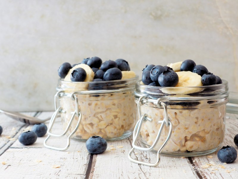 2 Gläser Overnight Oats garniert mit Blaubeeren und Banananscheiben, drumherum liegen einige Blaubeeren.