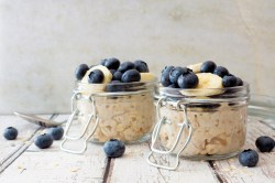 2 Gläser Overnight Oats garniert mit Blaubeeren und Banananscheiben, drumherum liegen einige Blaubeeren.