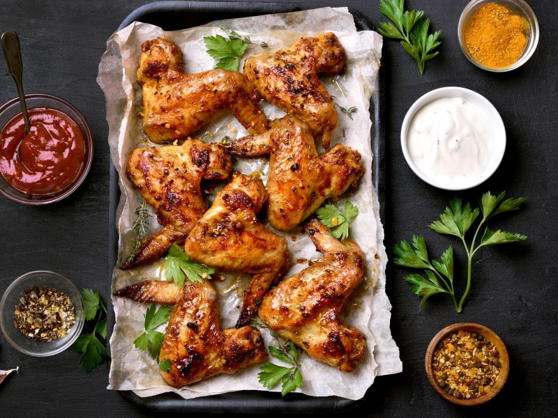 Honig Chicken Wings auf einem mit Backpapier ausgeschlagenen Backblech. Daneben stehen Schüsseln mit Soße.