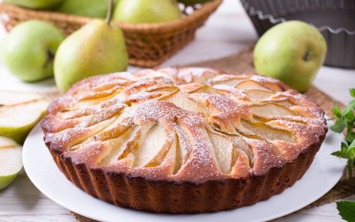 Ein Birnenkuchen mit Puderzucker auf einem Teller. Im Hintergrund liegen frische Birnen.