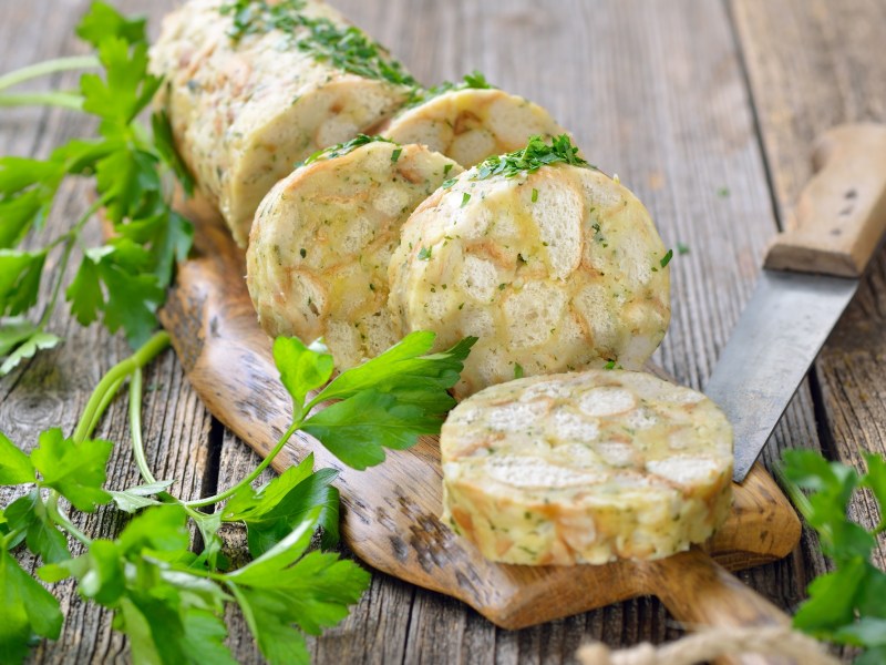 Ein angeschnittener Serviettenknödel auf einem Brett, daneben ein Messer und Petersilie.