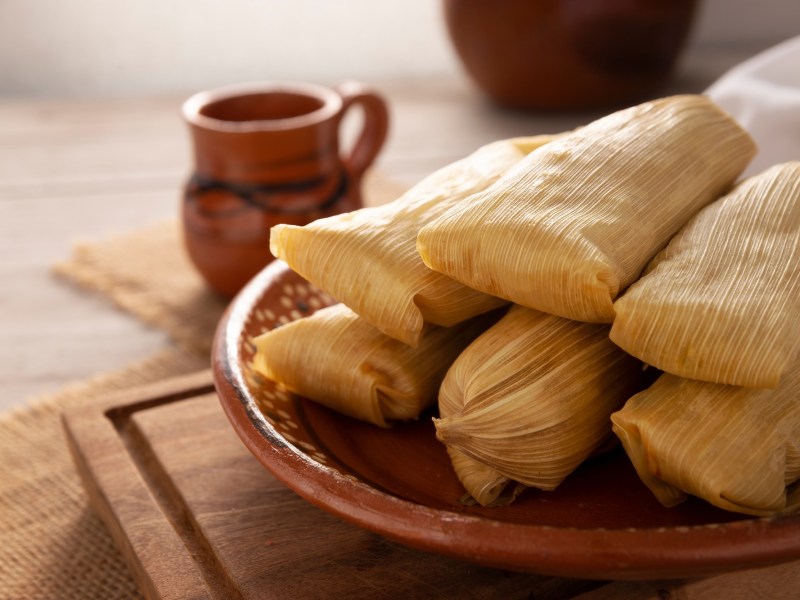 Tamales: mexikanische Mais-Teigtaschen auf einem Teller. Dahinter eine Tasse heiße Schokolade.