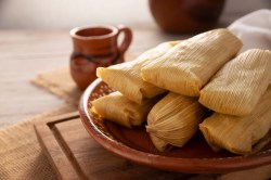 Tamales: mexikanische Mais-Teigtaschen auf einem Teller. Dahinter eine Tasse heiße Schokolade.