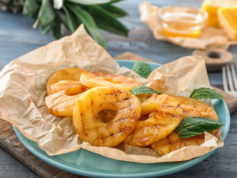 Ein hellblauer Teller ausgelegt mit Backpapier auf dem Ananasringe aus dem Airfryer liegen. Garniert sind die Ananasringe mit frischer Minze. Im Hintergrund steht eine kleine Schale mit Honig und die Blätter einer Ananas sind zu sehen.