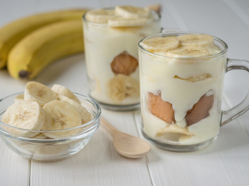 Zwei Dessertgläser mit Banana-Pudding. Daneben steht ein kleines Schälchen mit Bananenscheiben. Außerdem liegen zwei Bananen und ein Holzlöffel dabei.