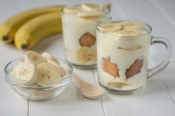 Zwei Dessertgläser mit Banana-Pudding. Daneben steht ein kleines Schälchen mit Bananenscheiben. Außerdem liegen zwei Bananen und ein Holzlöffel dabei.