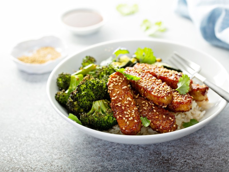 Brokkoli-Tempeh-Bowl mit geröstetem Brokkoli uns Reis,garniert mit Sesam. Eine Gabel liegt in der Schale.