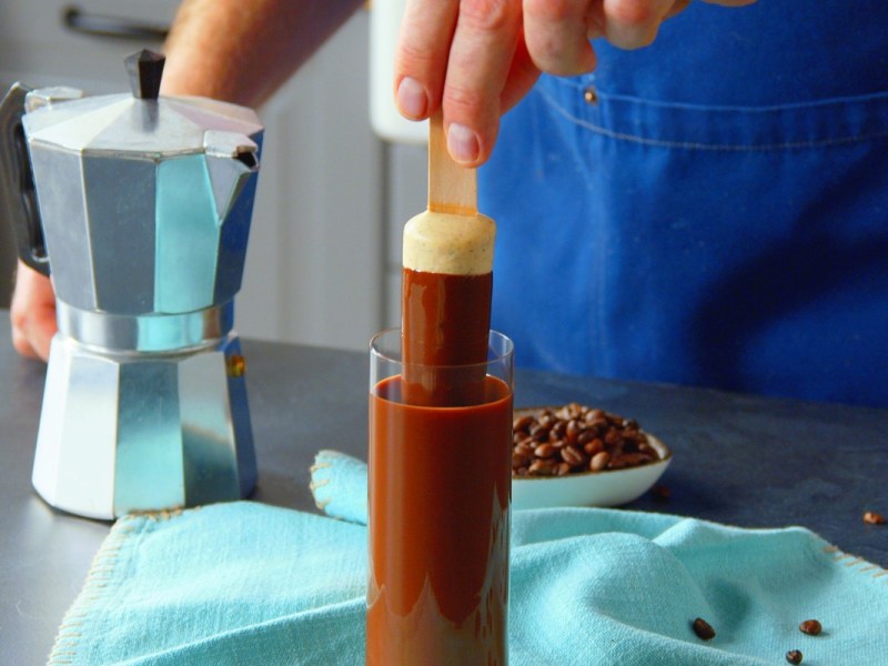 Ein Cheesecake am Stiel wird in ein Schnapsglas mit flüssigher Schokolade getaucht.