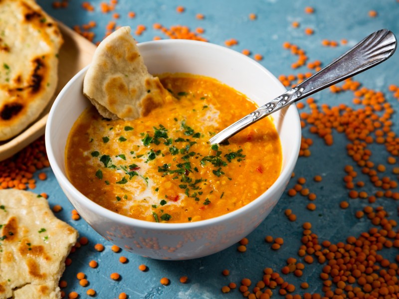 eine SchÃ¼ssel Curry-Linsen-Suppe mit einem StÃ¼ck Naanbrot und einem LÃ¶ffel. Daneben rohe rote Linsen und Brot.