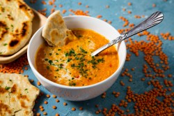 eine Schüssel Curry-Linsen-Suppe mit einem Stück Naanbrot und einem Löffel. Daneben rohe rote Linsen und Brot.