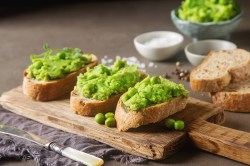 Erbsen-Basilikum-Hummus auf 3 Baguettescheiben, die auf einem Brett liegen. Im Hintergrund eine Schüssel mit dem Hummus und Brotscheiben.