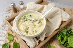 Eine Schüssel voll cremiger Gnocchi-Suppe mit Artischocken und Spinat serviert auf einem Holzbrett ausgelegt mit einer beigen Serviette. Daneben ist ein Salz- und Pfefferstreuer zu sehen. Außerdem steht eine kleine Schale mit Spinatblättern daneben.