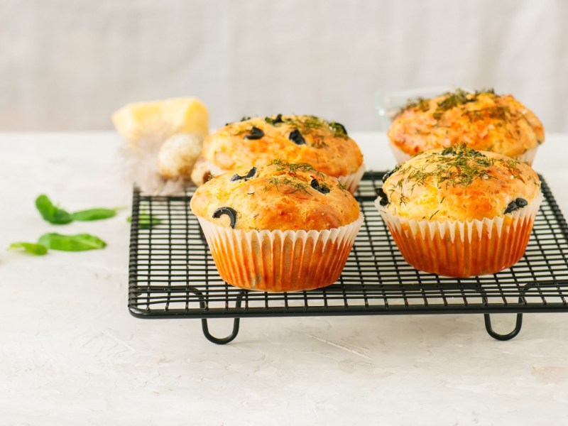 Herzhafte Haferfluckenmuffins mit Kräutern und schwarzen Olivenringen stehen auf einem schwarzen Gitter angerichtet. Im Hintergrund liegen ein paar frische Basilikumblätter.