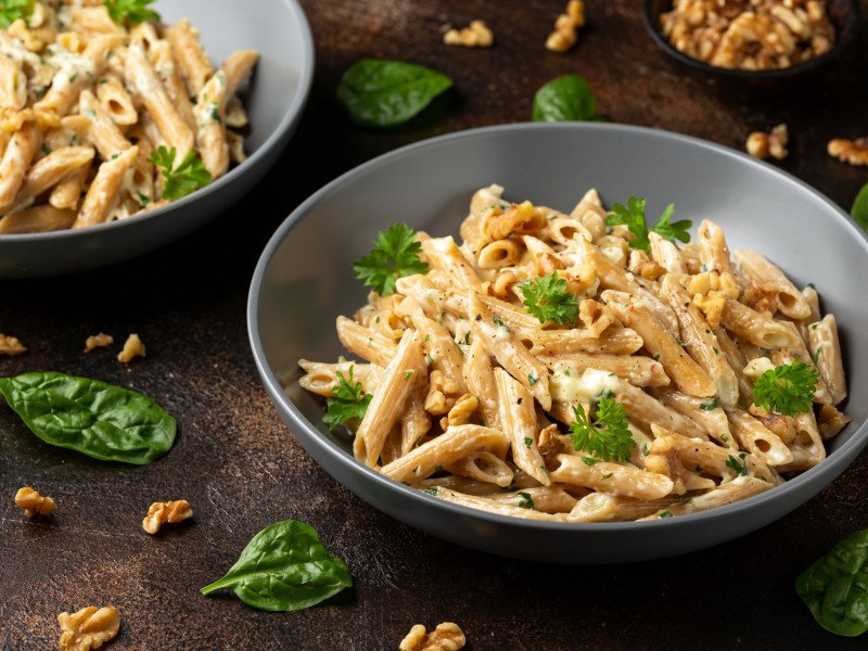 Ein grauer Teller mit einer großen Portion Hummus-Pasta, die mit frischer Petersilie und gehackten Walnüssen serviert wird. Im Hintergrund ist eine weitere Portion des Gerichts zu sehen. Auf dem Tisch liegen Basilikumblätter und Walnussstückchen verteilt.