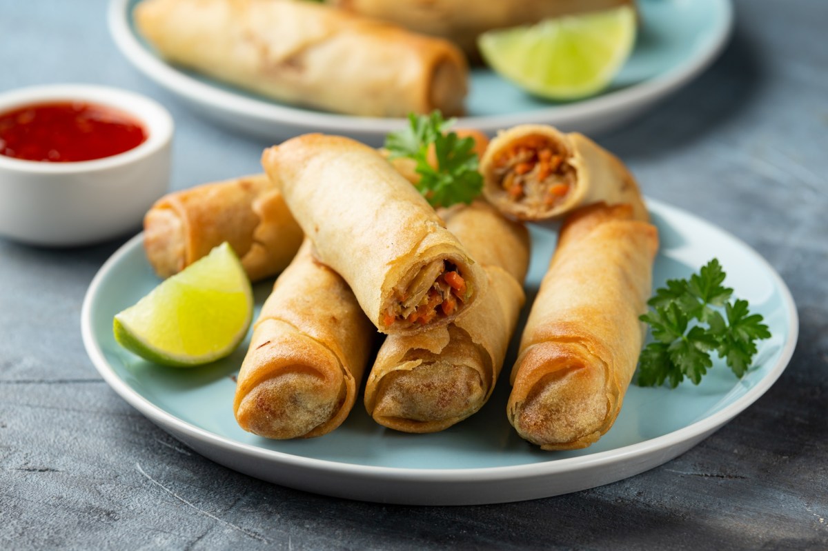 Kimchi-Frühlingsrollen liegen auf einem hellblauen Teller. Sie sind mit einem Limettenstück und frischer Petersilie garniert. Im Hintergrund steht eine Schale mit Soße, sowie ein weiterer Teller Frühlingsrollen.