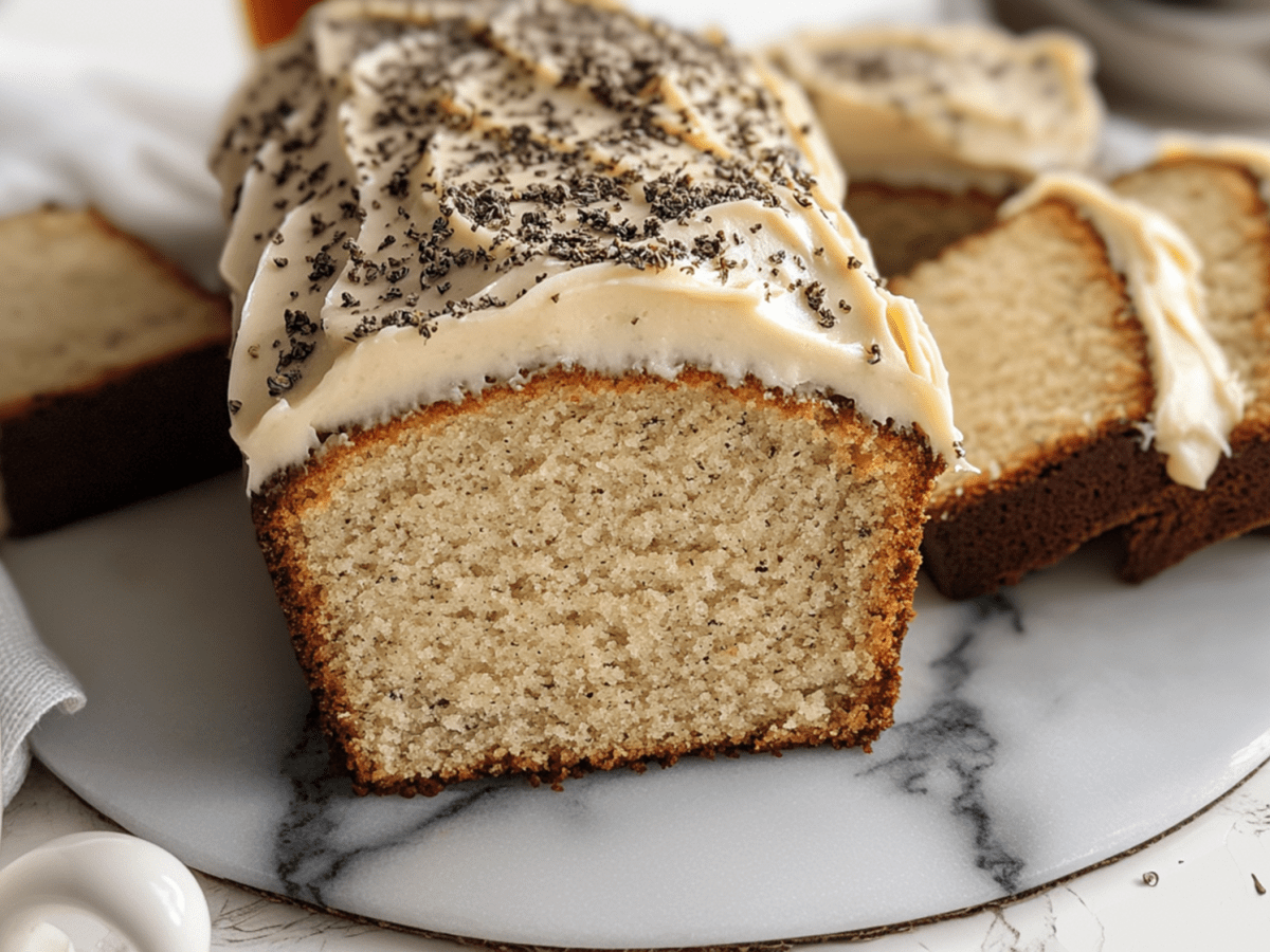 Ein London Fog Cake mit hellem Butterfrischkäse-Frosting und Earl Grey Tee bestreut. Der Kuchen ist kastenförmig und liegt angeschnitten auf einer Marmorplatte. Rechts daneben liegen ein paar weitere Scheiben London Fog Cake. Links auch eine Scheibe. Außerdem ist in der linken unteren Bildecke eine Tasse mit Earl Grey Teemischung zu sehen.