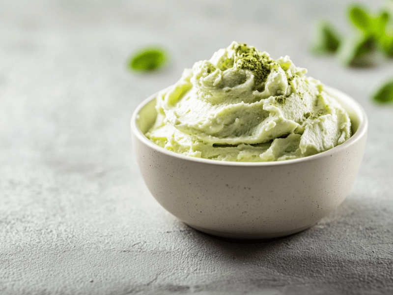 Eine kleine Schale mit cremigem Matcha-Aufstrich bestreut mit einer kleinen Prise Matcha-Pulver. Im Hintergrund sind verschwommen ein paar grüne Blättchen zu erkennen. Die Schale steht auf hellem Untergrund.