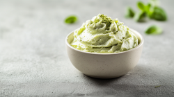 Eine kleine Schale mit cremigem Matcha-Aufstrich bestreut mit einer kleinen Prise Matcha-Pulver. Im Hintergrund sind verschwommen ein paar grüne Blättchen zu erkennen. Die Schale steht auf hellem Untergrund.