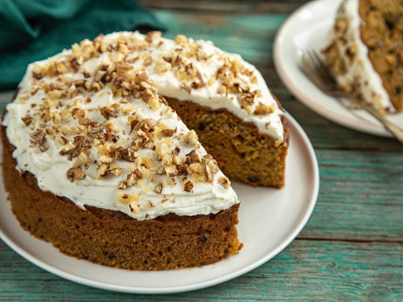 Ein runder Nusskuchen ohne Mehl steht auf einem weißen Teller auf einem grünlichen Holztisch. Er ist mit weißem Frosting versehen und mit Nussstückchen dekoriert. Im Hintergrund steht ein Stück des Kuchens.