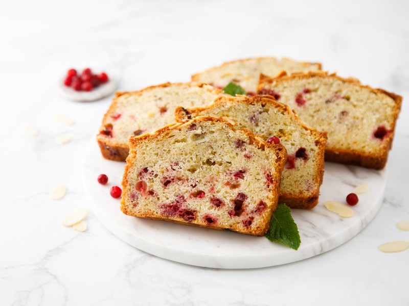 Eine heller marmorner Teller mit fünf Scheiben Preiselbeerkuchen. Als Garnitur liegen frische Minzblätter dabei. Ringsum liegen einige Mandelsplitter und frische Preisebeeren. Im Hintergrund ist ein kleines Schälchen mit Preiselbeeren zu sehen.