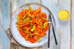 Ein Teller mit Räucherlachs-Carpaccio. Daneben ist ein kleines Schälchen Dressing. Das Ganze steht auf einem hellblauen Hintergrund.