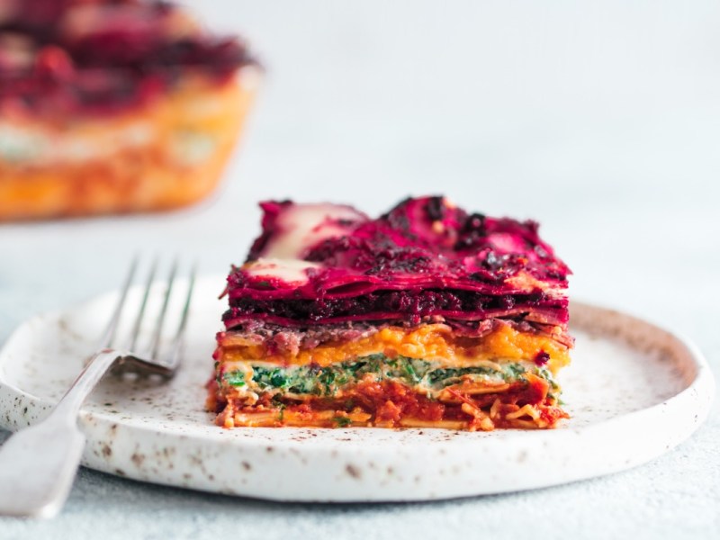 Ein Stück vegane Regenbogen-Lasagne auf einem Teller. Der Rest ist im Hintergrund zu sehen.