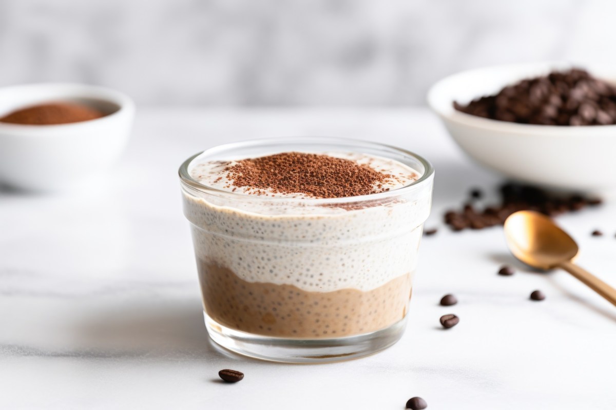 Eine Glasschale mit Tiramisu-Chia-Pudding, bestehend aus einer dunklen Kaffee-Schokoschicht und einer hellen Vanilleschicht steht auf einer marmornen Arbeitsfläche. Der Tiramisu-Chia-Pudding ist mit Kakaopulver bestreut. Im Hintergrund steht eine weiße Schale mit Kaffeebohnen und eine kleine Schale mit Kakaopulver. Außerdem sind ein paar Kaffeebohnen verstreut.