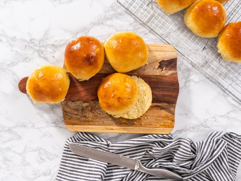 vegane Brioche-Brötchen auf einem Brett, teilweise aufgeschnitten, daneben ein paar weitere Brötchen auf einem Kuchengitter. Draufsicht.