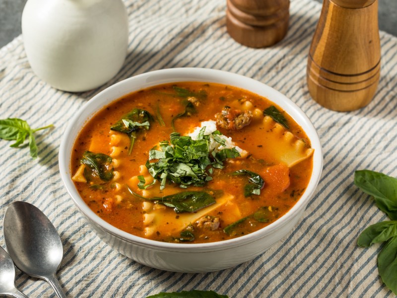 Lasagnesuppe in einer weißen Schüssel steht auf einer gesteiften Tischdecke. Drumherum liegen ein Löffel, Basilikumblätter sowie ein Pfeffer- und Salzstreuer. Die Suppe ist ebenfalls mit Basilikum und Käse garniert.