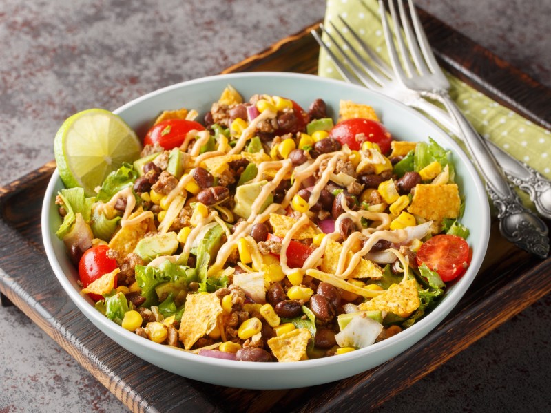 Bunter und veganer Nacho-Salat in einer weißen Schüssel auf einem dunklen Holzbrett. Der Salat besteht aus Nachos, Kidneybohnen, Nachos, frischem Salat, Mais und Tomaten. Links neben der Schale liegen zwei Gabeln auf einer Serviette.