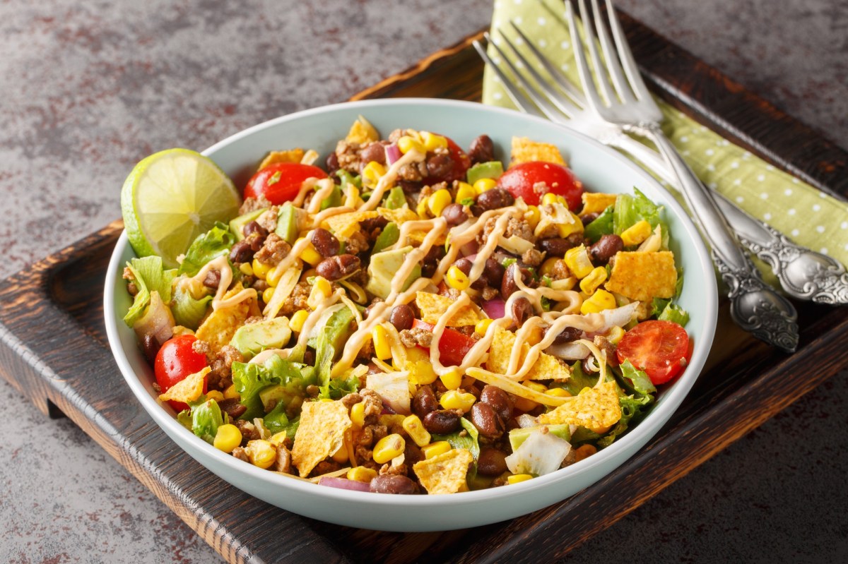 Bunter und veganer Nacho-Salat in einer weißen Schüssel auf einem dunklen Holzbrett. Der Salat besteht aus Nachos, Kidneybohnen, Nachos, frischem Salat, Mais und Tomaten. Links neben der Schale liegen zwei Gabeln auf einer Serviette.