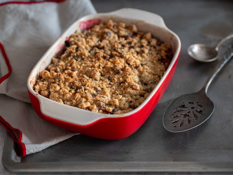 Veganer Porridgeauflauf in einer weiß-roten Auflaufform. Man sieht am Rand, dass er mit Beeren zubereitet ist. Links neben ihm liegt ein weiß-rotes Tuch, rechts zwei Löffel.