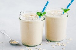 Zwei Gläser Käsekuchen-Shake mit blauen Strohhalmen und frischer Minze dekoriert. Die Käsekuchen-Shakes sind jeweils mit ein paar Haferflocken bestreut. Neben den Gläsern liegt ein Löffel mit Haferflocken und ringsum sind auch welche verstreut. Die Gläser stehen auf einer weißen Holzoberfläche.