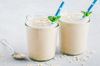 Zwei Gläser Käsekuchen-Shake mit blauen Strohhalmen und frischer Minze dekoriert. Die Käsekuchen-Shakes sind jeweils mit ein paar Haferflocken bestreut. Neben den Gläsern liegt ein Löffel mit Haferflocken und ringsum sind auch welche verstreut. Die Gläser stehen auf einer weißen Holzoberfläche.