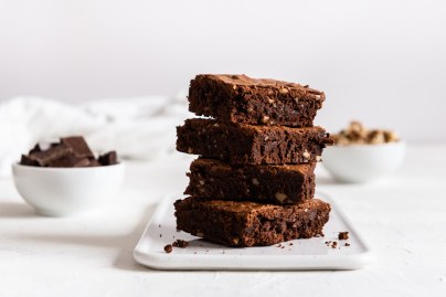 Ein Stapel zuckerfreie Apfel-Brownies auf einem weißen, eckigen Teller. Im Hintergrund stehen eine Schüssel Schokolade und eine mit Walnüssen.