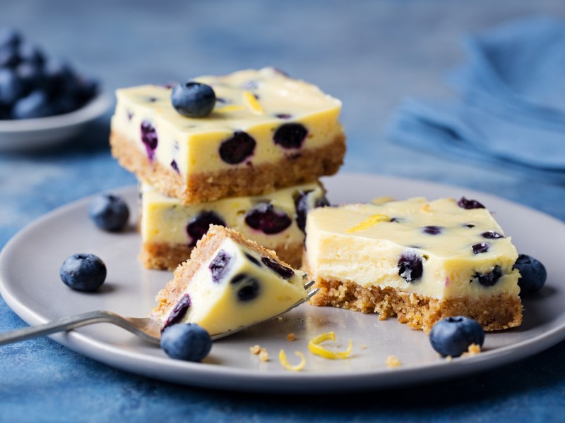 Mehrere Stücke zuckerfreier Zitronen-Heidelbeer-Kuchen auf einem Teller. Drum herum liegen Blaubeeren.