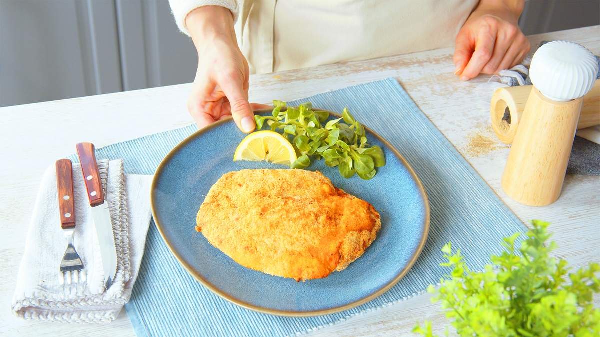 Ein Cordon Bleu mit Salami auf einem blauen Teller. Auf dem Teller liegt eine Zitronenspalte und etwas Feldsalat. Neben dem Teller liegt etwas Besteck und eine Salz und Pfeffermühle.