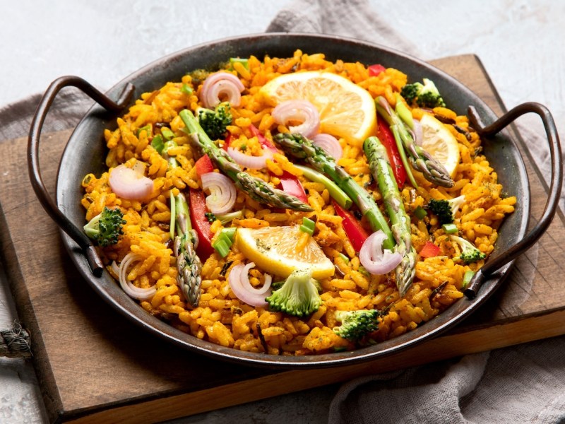 Mediterrane Reispfanne mit Spargel, Brokkoli, Paprika und Zwiebel in einer gusseisernen Pfanne, die auf einem Holzbrett steht. Nahaufnahme.