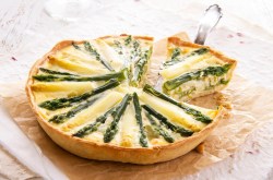 Eine runde, angeschnittene vegane Spargelquiche, aus der ein Stück herausgenommen wird, auf einem Stück Backpapier.