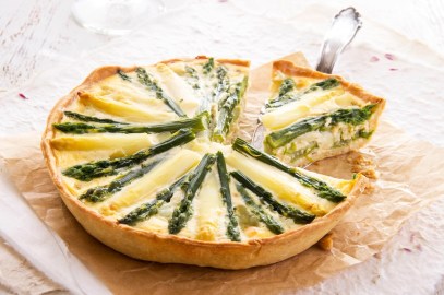 Eine runde, angeschnittene vegane Spargelquiche, aus der ein Stück herausgenommen wird, auf einem Stück Backpapier.